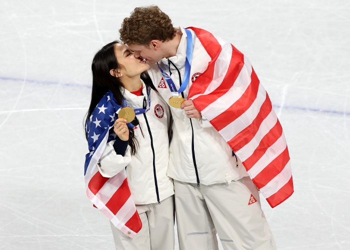 Two Olympic athletes wrapped in American flags sharing a moment on ice, highlighting the Olympic Village atmosphere.