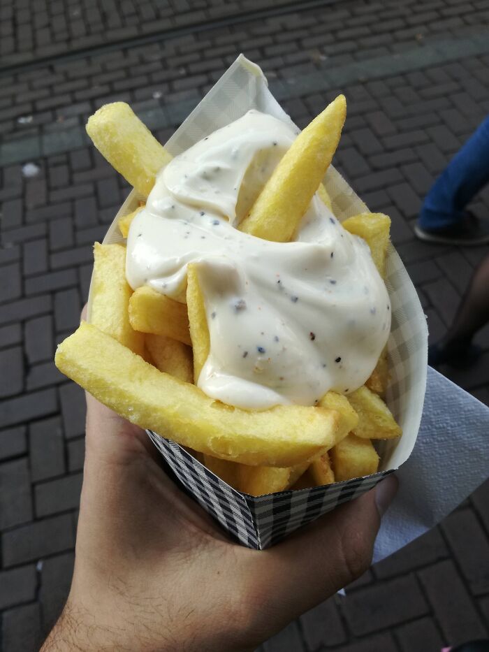 Hand holding seasoned fries with creamy white sauce on a city street, showing things people have in their country.