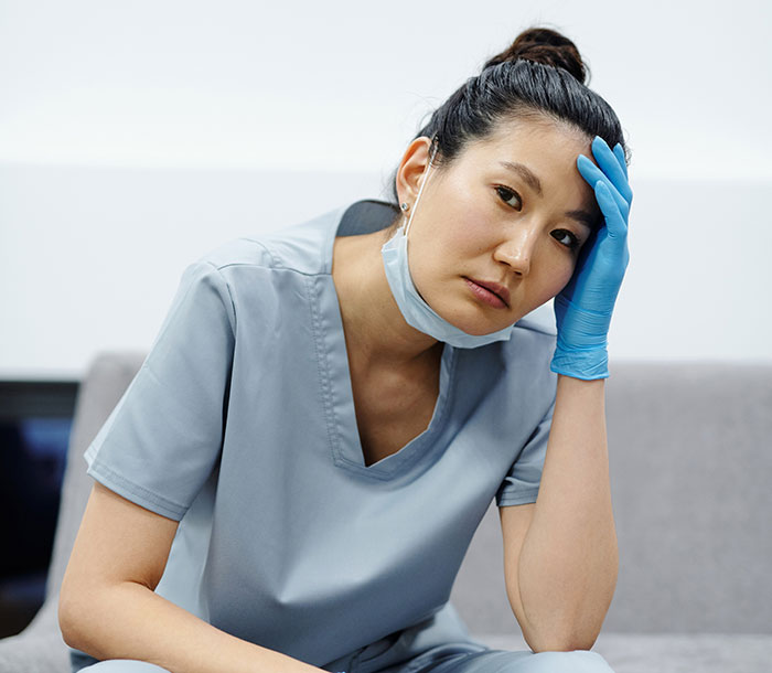 Stressed nurse wearing scrubs and gloves sitting with hand on forehead, illustrating patients helping doctors and nurses.