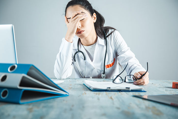 A stressed female doctor sitting at a desk with medical files and stethoscope, showing fatigue from patient care demands.