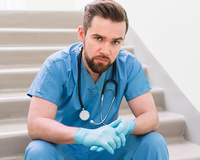 Male doctor in blue scrubs and gloves sitting on stairs, reflecting on patients’ actions affecting doctors and nurses.