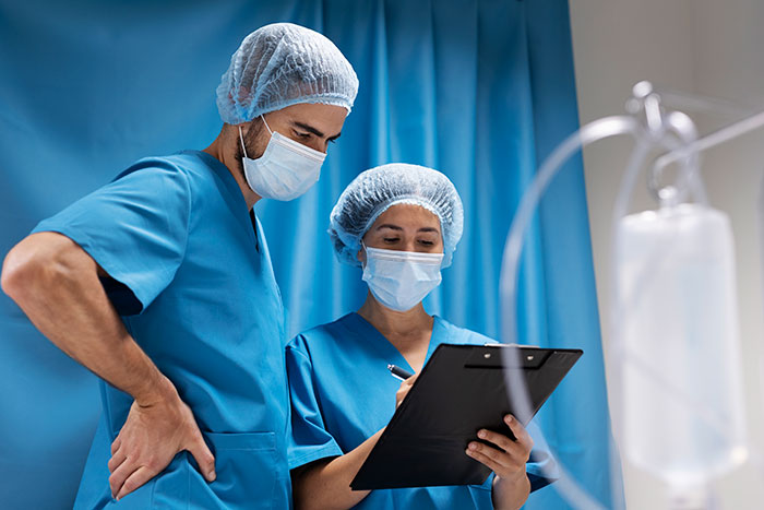 Two medical professionals in scrubs and masks reviewing patient notes in a clinical setting about patient behaviors.