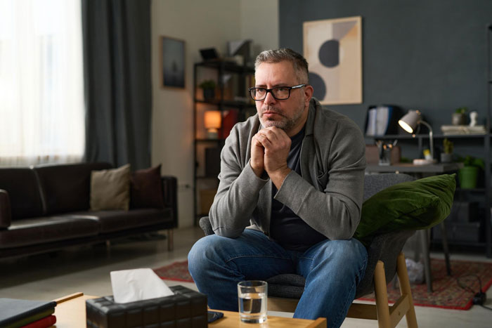 Middle-aged man in casual clothes sitting thoughtfully in living room, reflecting on woman's easy-going lifestyle and affair discovery.
