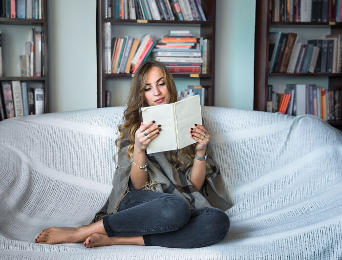 Young woman reading a book on a sofa in a cozy room filled with books, reflecting on petty things people judge others for.