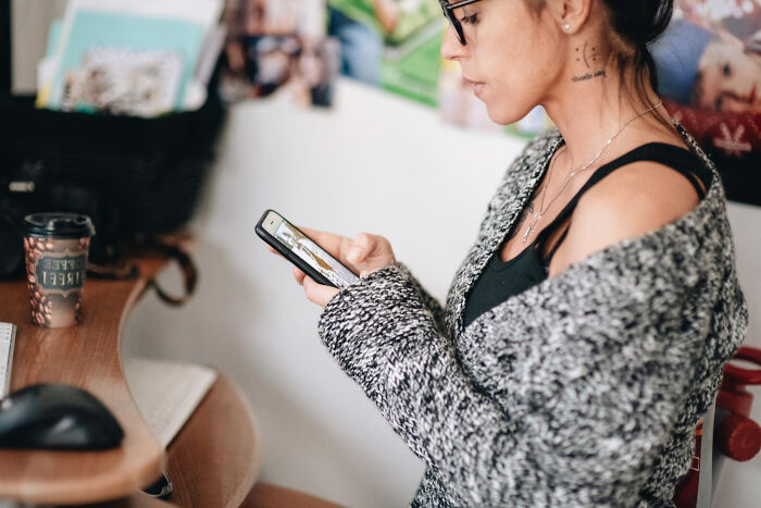 Person in glasses and a gray sweater secretly judging others while looking at their phone indoors, illustrating petty things people judge.