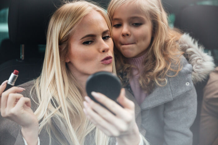 Woman applying lipstick while a young girl watches inside a car, capturing surprisingly petty things people secretly judge others for.