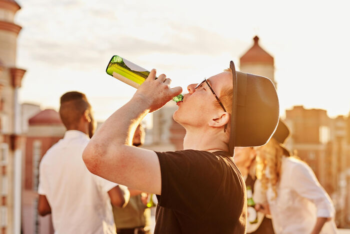Young man wearing glasses and a hat drinking from a bottle at a rooftop party, highlighting petty things people secretly judge.