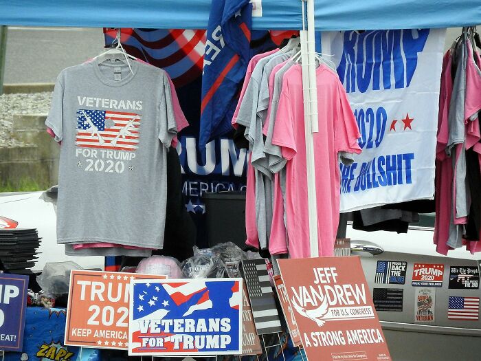 Outdoor political merchandise booth displaying Veterans for Trump 2020 shirts and campaign signs, highlighting petty judgments.