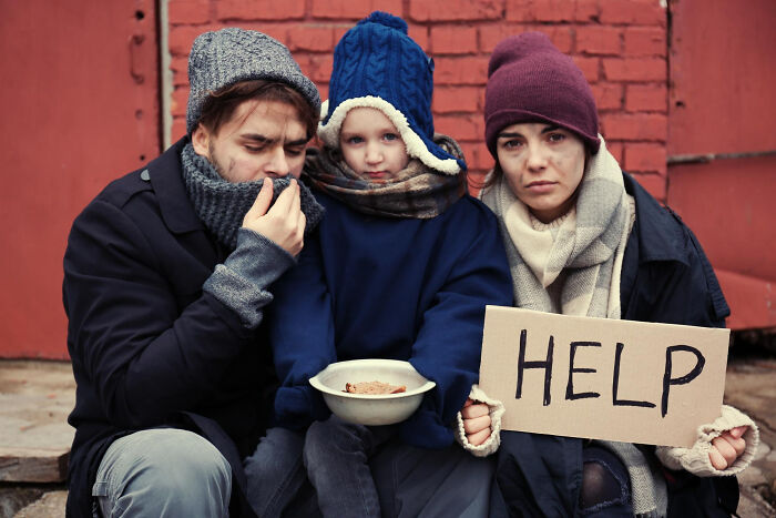 A family sitting outside against a brick wall, holding a help sign, illustrating petty judgments people secretly make.