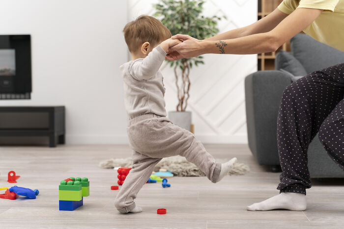 Toddler learning to walk with help in a messy room scattered with toys, illustrating uncomfortable and messy secrets concept.