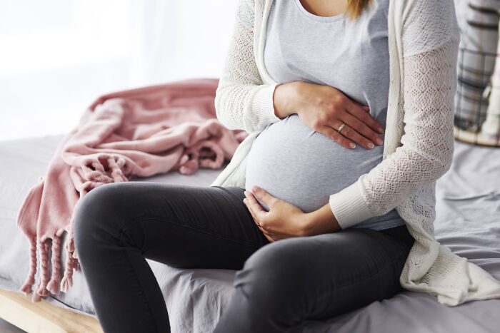 Pregnant woman sitting on bed, holding belly, illustrating uncomfortable and messy secrets confessed anonymously.