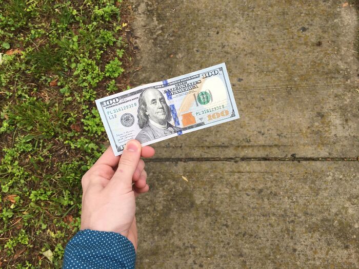 Hand holding a one hundred dollar bill outdoors on a concrete sidewalk near grass, symbolizing messy secrets about money.