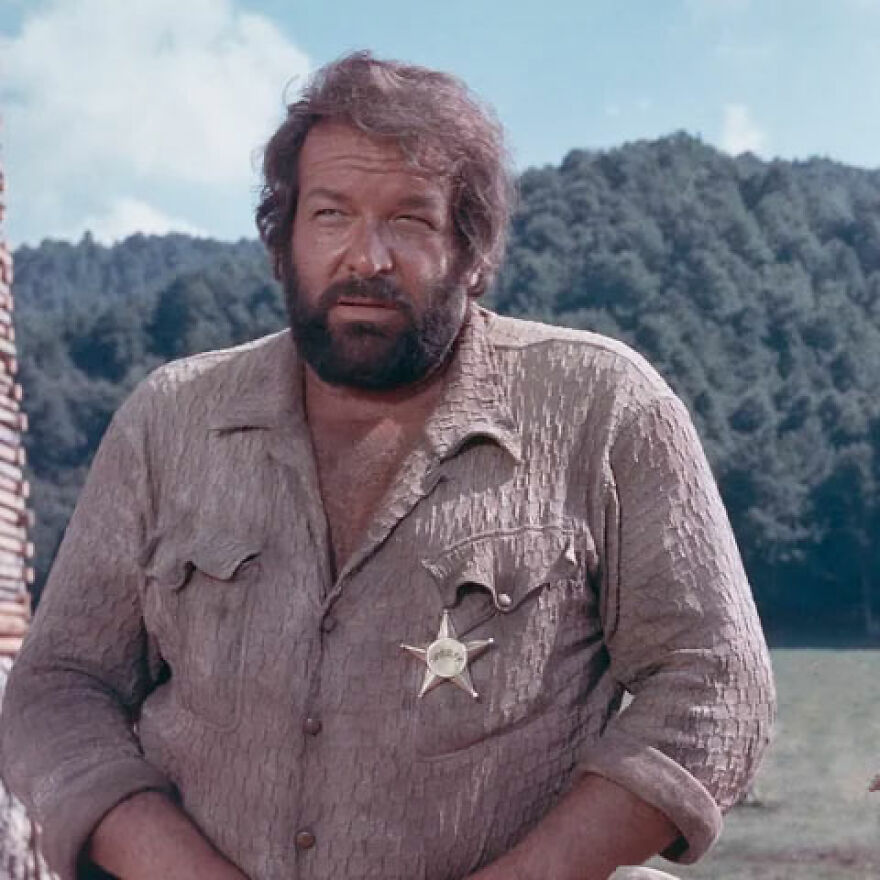 Bearded man wearing a brown shirt with a sheriff badge, standing outdoors with mountains and trees in the background representing gigachads.