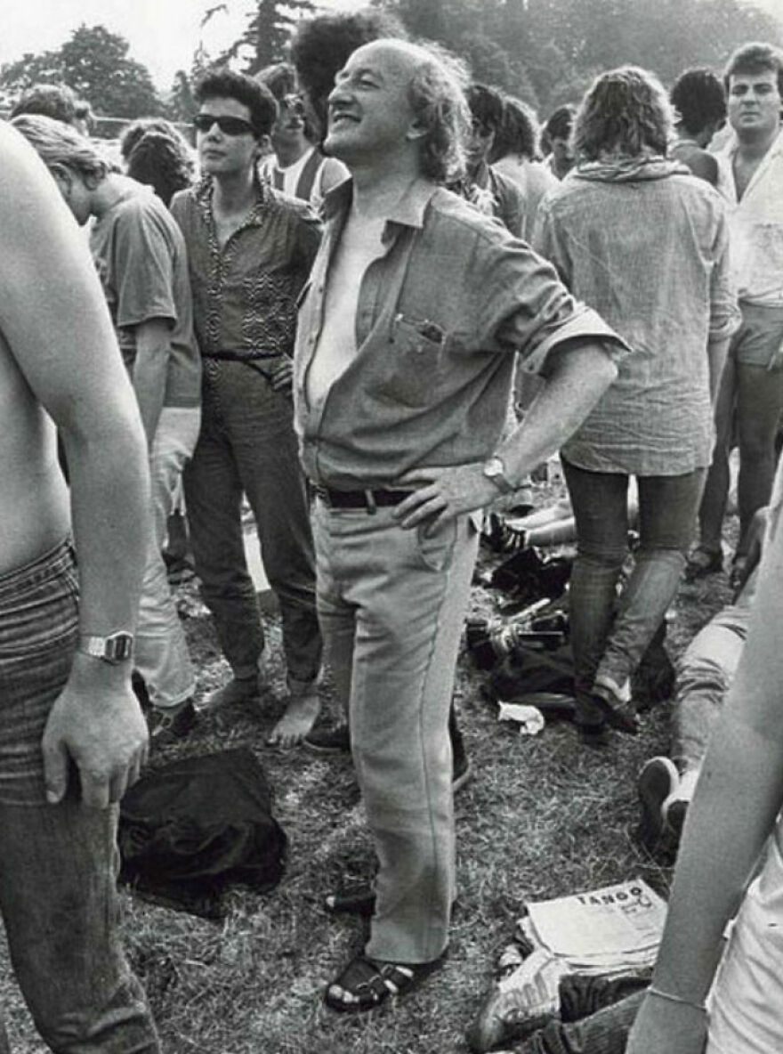 Older man confidently standing among a crowd outdoors, wearing sandals and casual clothing, embodying gigachads admiration.