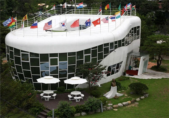 Unique architectural malpractice building with irregular windows and flags on roof, surrounded by gardens and outdoor seating.