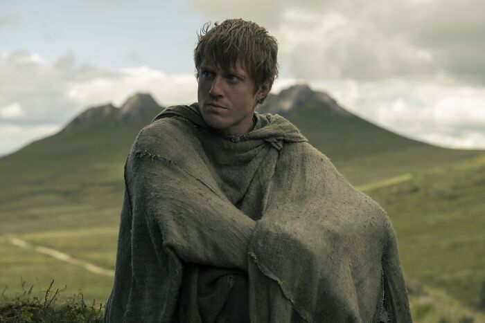 Character from A Knight of the Seven Kingdoms wearing a cloak, set against a mountainous background in a medieval landscape.