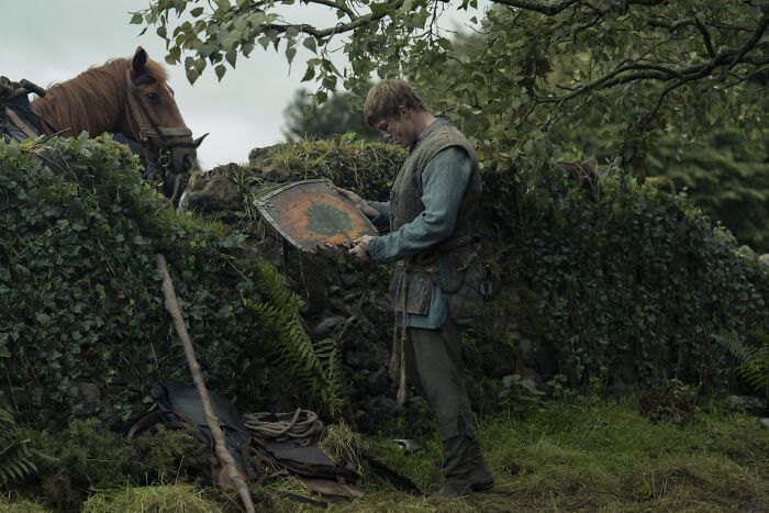 A Knight Of The Seven Kingdoms season 2 scene with a medieval character examining a shield near a horse in a forest.