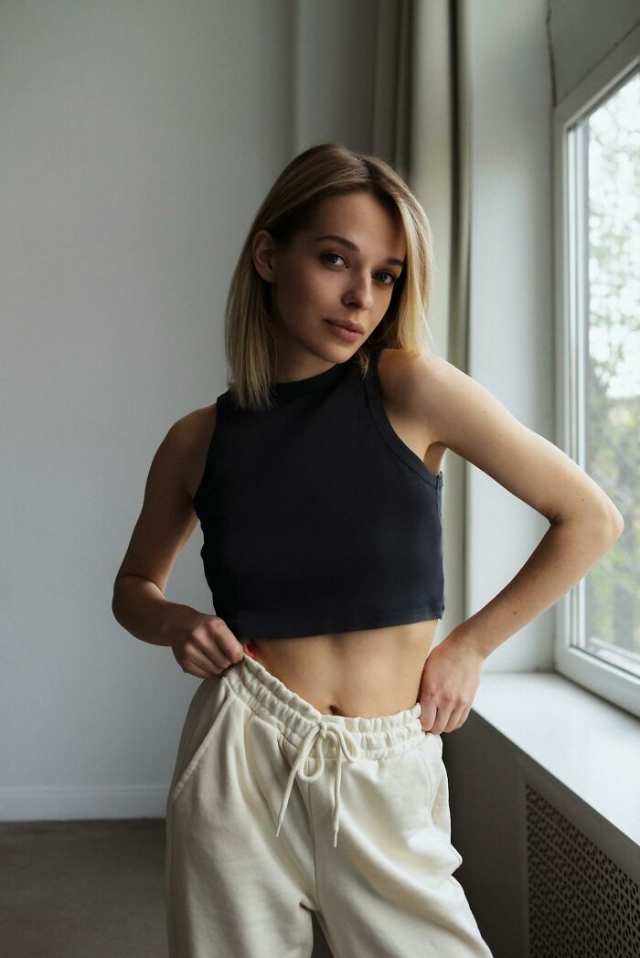 Young woman in a black crop top and white sweatpants standing by a window, representing toxic ways women traumatized men.
