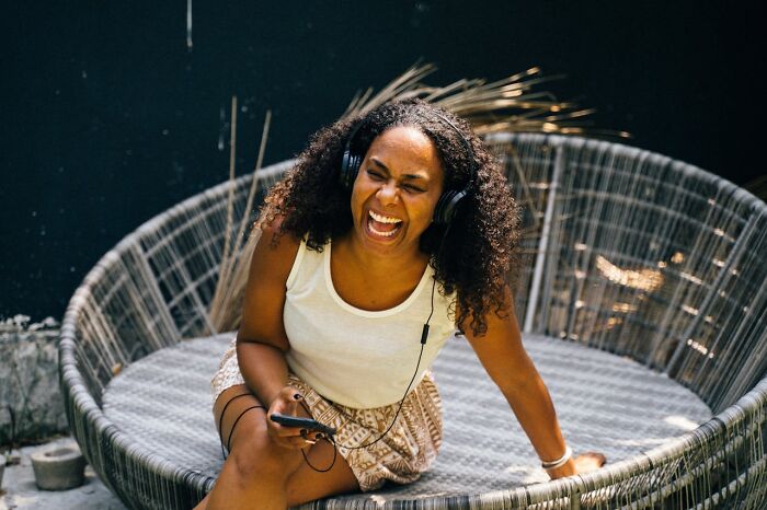 Woman with headphones laughing joyfully while sitting on a woven lounge chair, reflecting unhinged toxic ways women traumatized men.