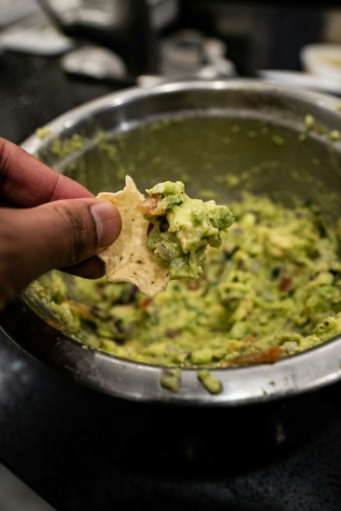 Hand holding a tortilla chip with guacamole, focusing on the avocado dip in a metal bowl, unhinged toxic ways keyword concept.