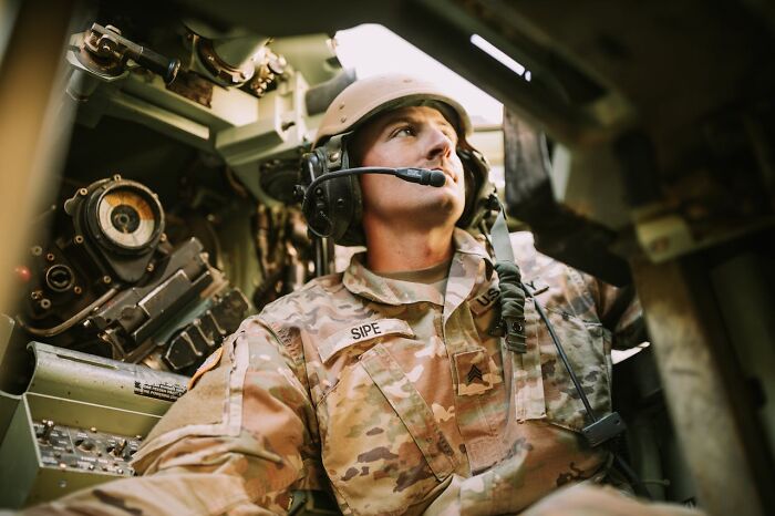 Soldier in military uniform inside armored vehicle, wearing headset and communicating amid sounds signaling immediate disaster.