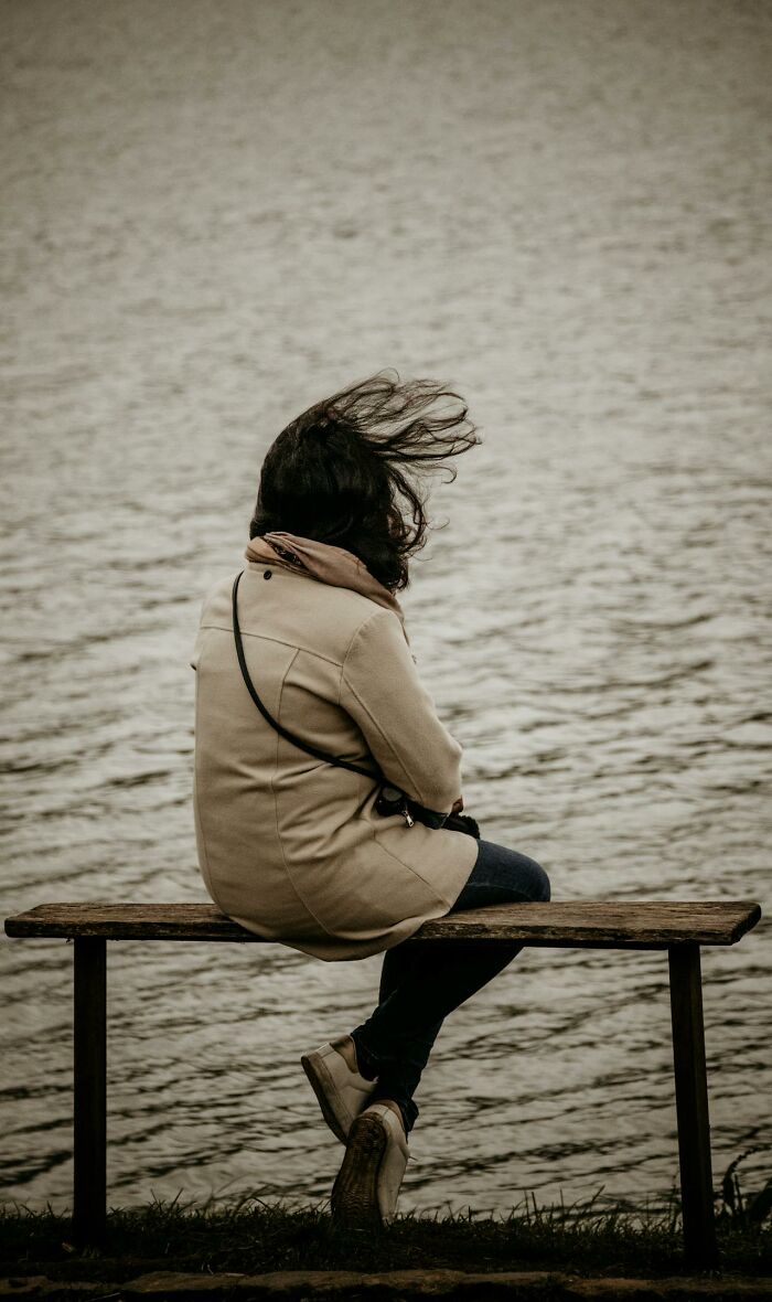 Person sitting alone by the water with hair blowing in the wind, reflecting on silent judgment from others doing 97 things