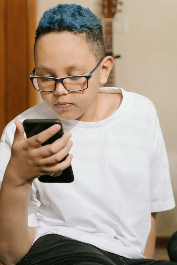 Young boy wearing glasses and a white shirt, looking intently at smartphone, highlighting people silently judge others concept.