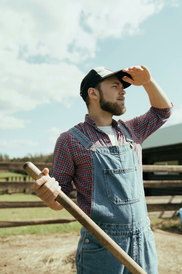 Young man in denim overalls and cap holding a wooden tool, looking into distance with a thoughtful pause outdoors.