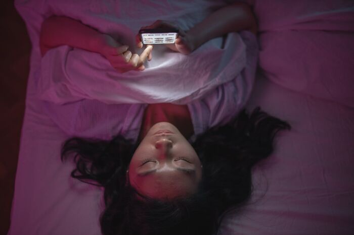 Person lying in bed under soft purple light, looking at phone screen, illustrating funny and harmless conspiracy theories concept.