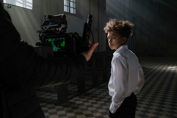 Young actor in white shirt on film set, being recorded by a camera operator in a dimly lit room with checkered floor.