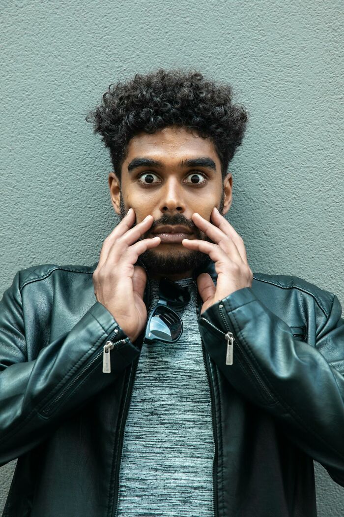 A man with curly hair and wide eyes expressing fear, wearing a black leather jacket against a textured gray wall.
