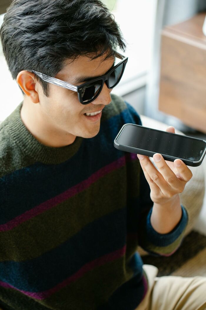 Young man wearing sunglasses holding phone, smiling while using voice assistant to find today I learned facts.