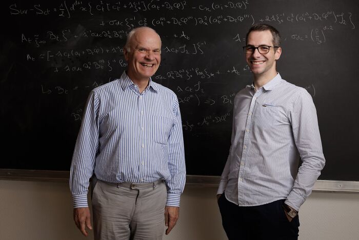 Two men smiling and standing in front of a chalkboard filled with complex equations, illustrating moments of stupid things paused for.