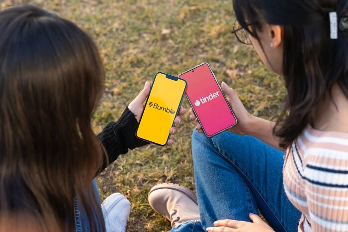 Two women sitting outdoors comparing dating apps on phones, a lighthearted moment amid funny harmless conspiracy theories.