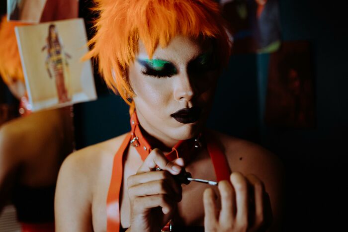 Person with vibrant orange hair and bold makeup focused on applying nail polish, related to narcissist quiz concept. Person with vibrant orange hair and bold makeup focused on applying nail polish, related to narcissist quiz concept.