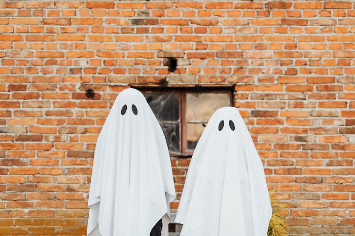 Two people dressed as ghosts with white sheets and black eye holes standing in front of a brick wall for funny harmless conspiracy theories.