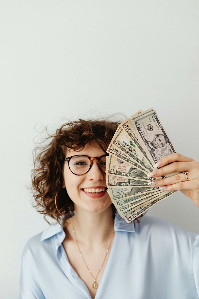 Young smiling woman holding cash fanned out near her face, illustrating people silently judge others with money.