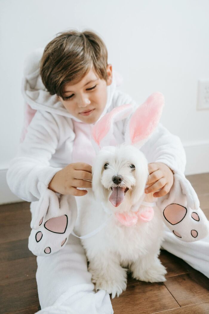 Child in a bunny costume gently holding a small white dog dressed with pink bunny ears and bow, showing silent judgment behavior.