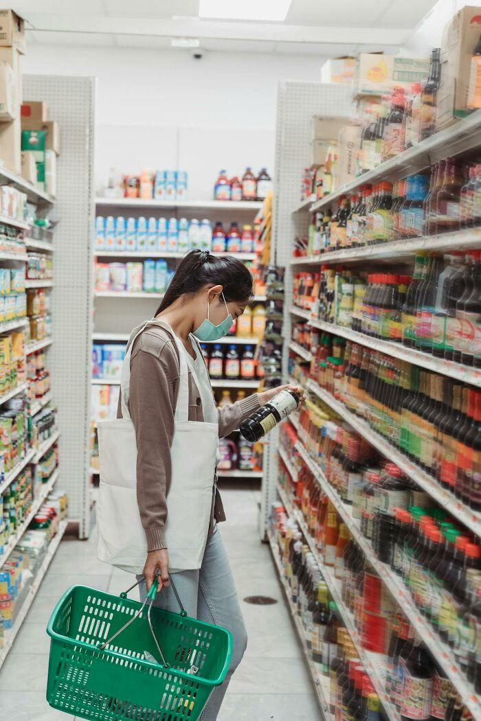 Woman wearing a mask shopping in grocery store aisle, examining a bottle among various products, funny conspiracy theories concept.