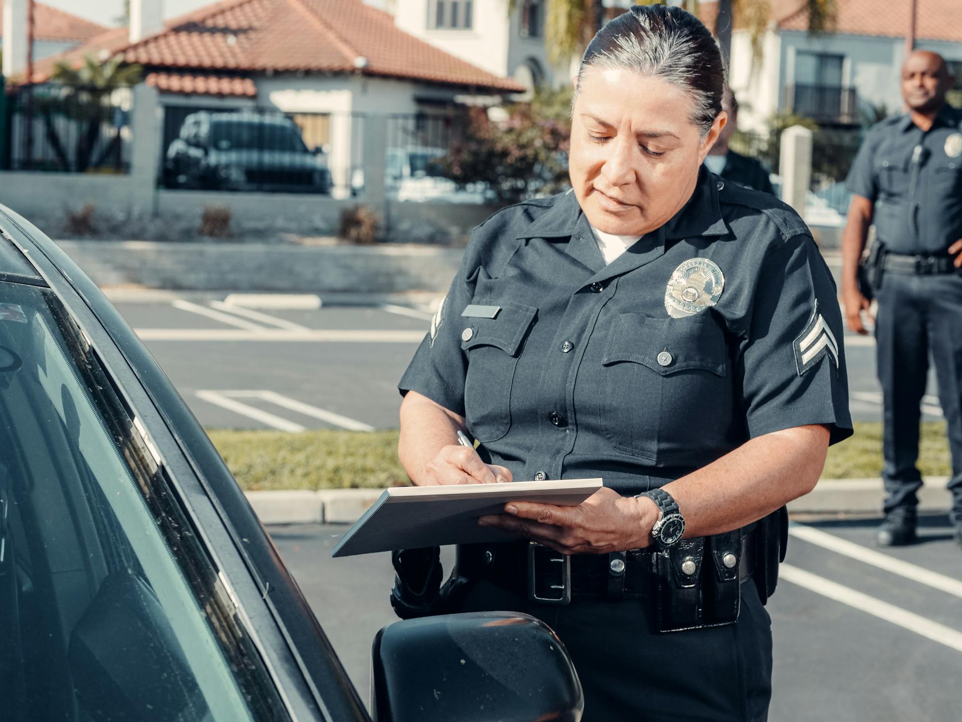 Female police officer writing a ticket near a car in a parking lot with a colleague standing in the background