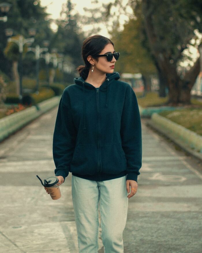 Woman in sunglasses and dark hoodie walking outdoors holding a drink, illustrating silent judgment of others behavior.
