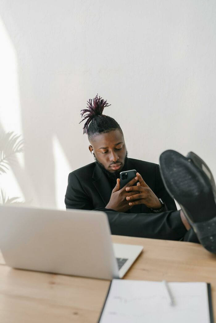 Man in black suit silently judging others while using phone, seated with feet up near laptop in bright office space