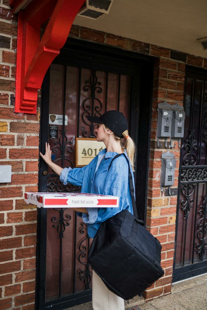 Young woman holding pizza boxes and delivery bag, ringing doorbell at a residential brick building entrance.