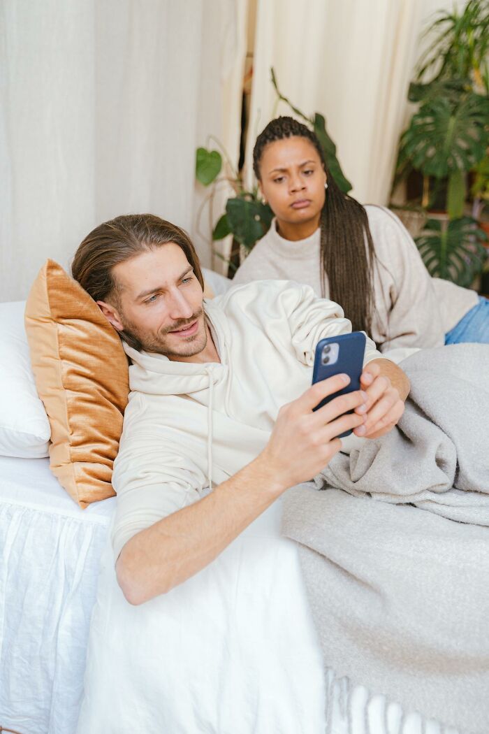 Couple in bed with woman looking upset while man is distracted by his phone, illustrating toxic ways women traumatized men.