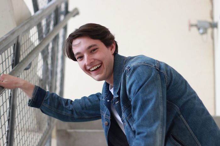 Young man in a denim jacket smiling and leaning on a metal railing, capturing a moment of surprised or stupid things said.