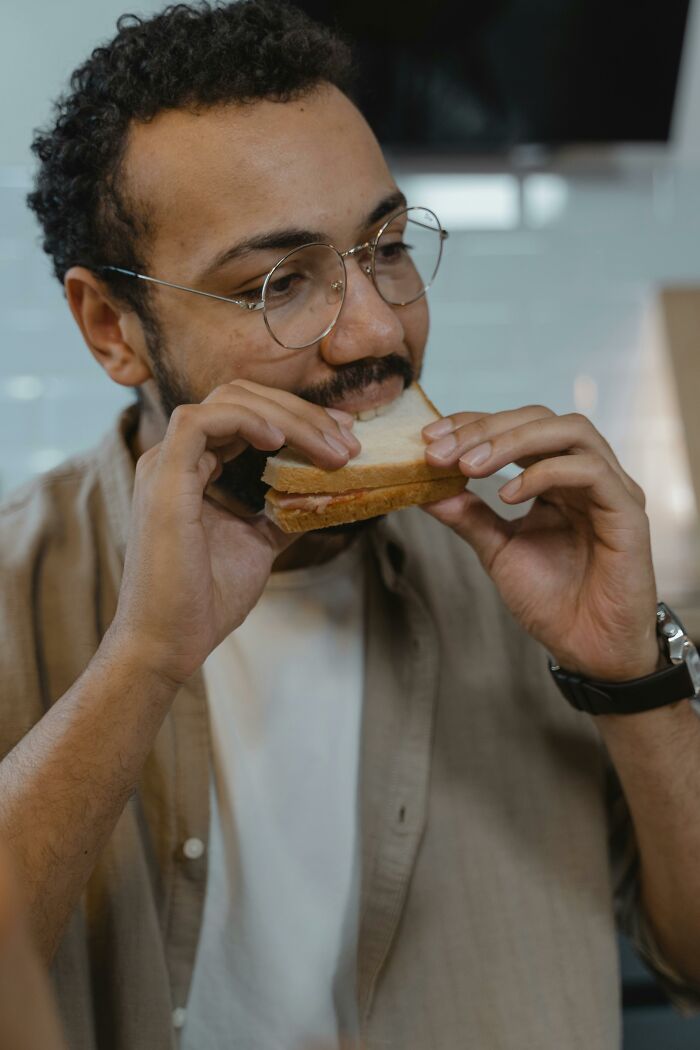 Man with glasses eating a sandwich indoors, representing men reflecting on unhinged and toxic ways they were traumatized by women.