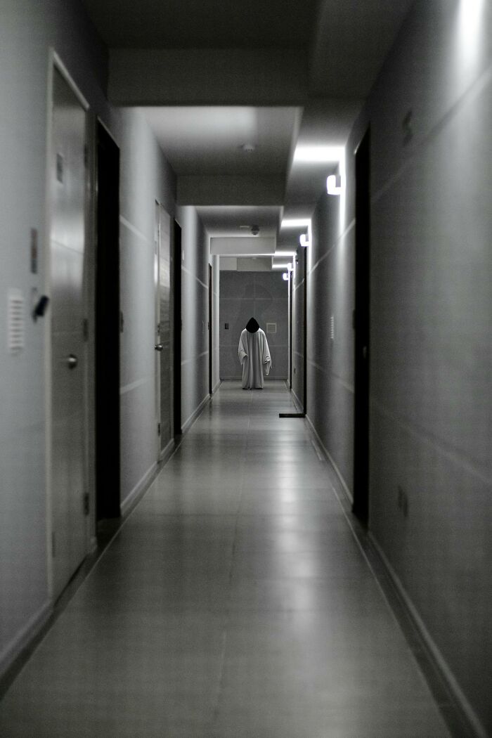Figure in white cloak standing alone at end of dimly lit hallway, evoking graveyard shift workers scared or confused.