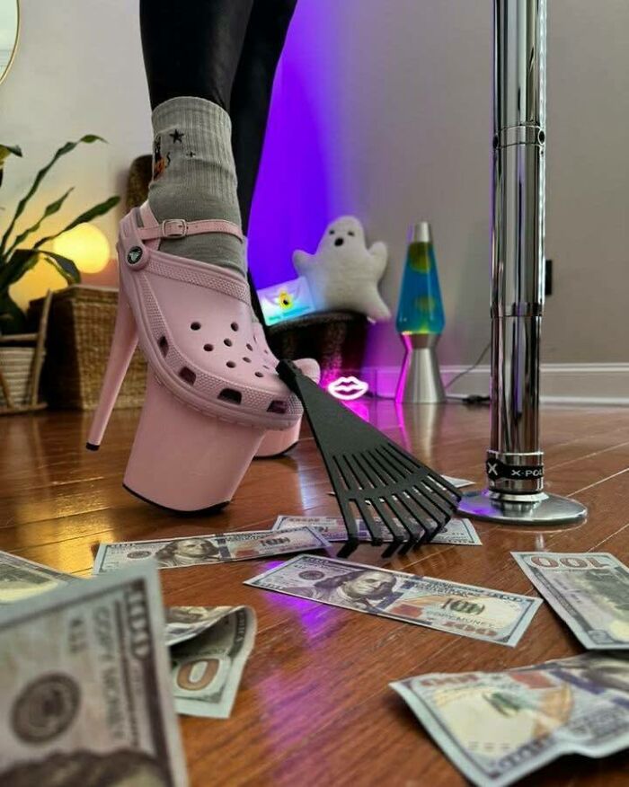 Person wearing pink high heel platform Crocs and socks, using a rake to gather scattered money on wooden floor, funny meme concept.