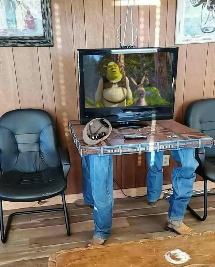 Television on a jeans table with legs wearing boots between black chairs showing an animated movie scene funny meme.