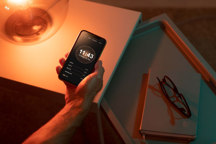 Hand holding phone with pill reminder app open beside glasses and books on a bedside table at night. Hand holding phone with pill reminder app open beside glasses and books on a bedside table at night.